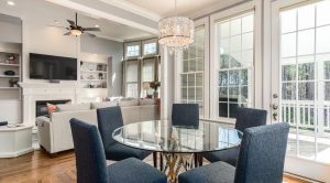 Modern open-concept living and dining area featuring a glass-top table with navy upholstered chairs, a cozy sitting area with a fireplace and mounted TV, large windows, and elegant natural lighting.